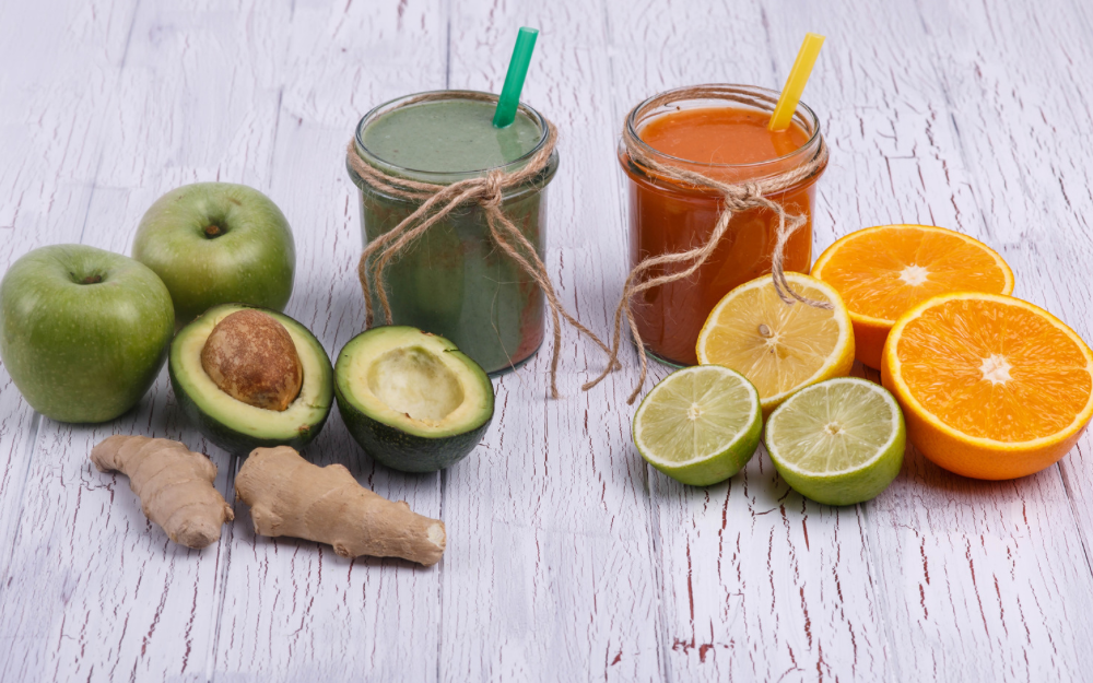 Fresh detox ingredients including green apples, avocado, ginger, limes, oranges, and two colorful smoothies in glass jars with straws on a rustic white wooden table—symbolizing natural metabolism and detox support through whole foods.