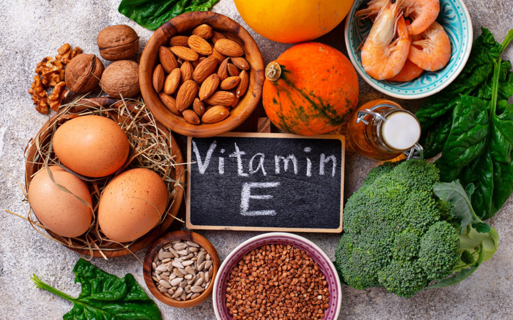 A colorful assortment of Vitamin E-rich foods, including eggs, almonds, walnuts, sunflower seeds, spinach, broccoli, shrimp, and pumpkin, arranged around a chalkboard sign labeled “Vitamin E”