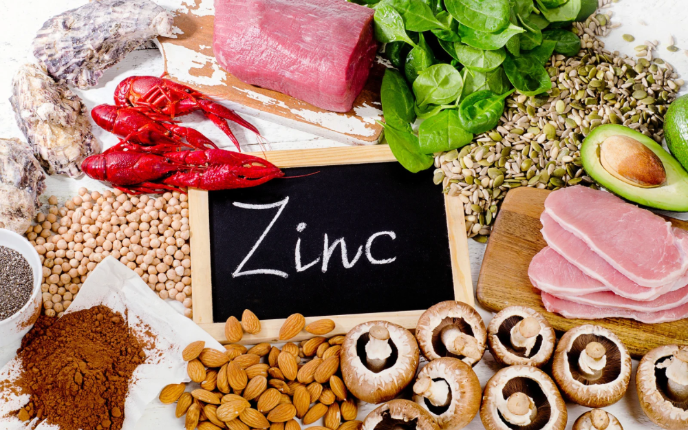 A variety of zinc-rich foods—including oysters, red meat, spinach, seeds, avocado, mushrooms, nuts, and legumes—surround a chalkboard labeled “Zinc,” highlighting natural sources that support immune and gut health.