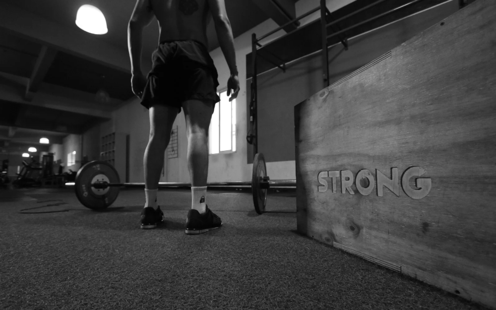 Black and white image of a man in a gym preparing to lift a barbell, with a wooden box labeled “STRONG” in the foreground—symbolizing strength training, fitness goals, and muscle-building supported by creatine supplements.