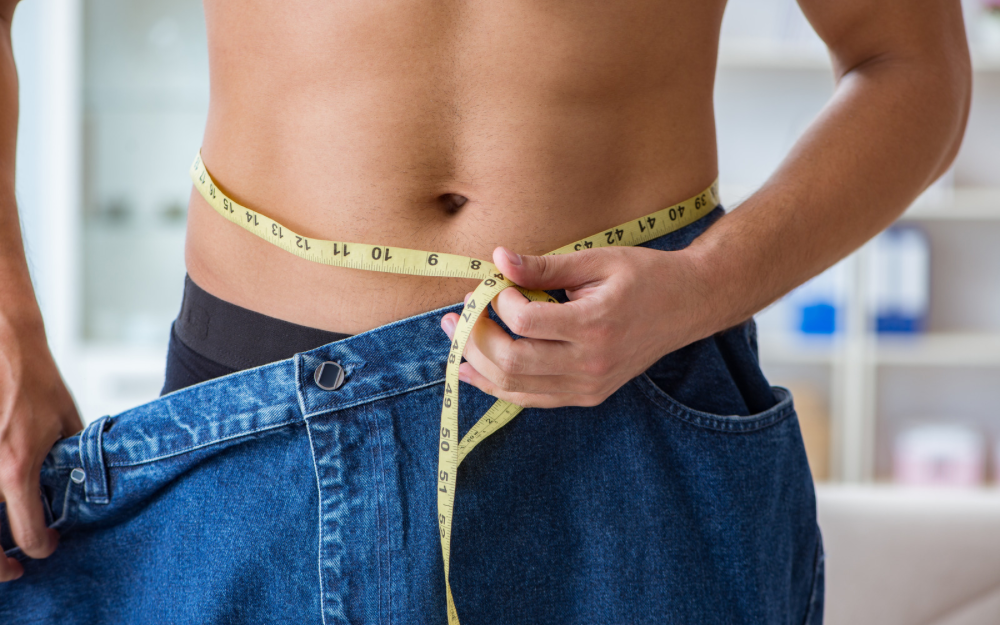 A person measures their waist with a tape measure while wearing oversized jeans, symbolizing fat loss and improved metabolism.