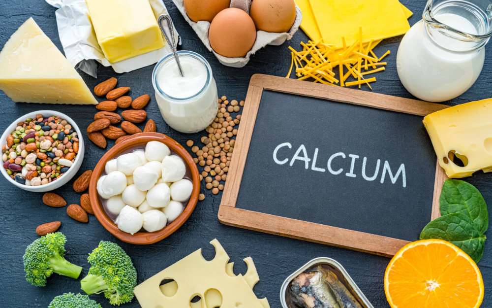 A variety of calcium-rich foods—including dairy, leafy greens, legumes, nuts, fish, and citrus—surround a chalkboard labeled “CALCIUM,” highlighting natural sources that support muscle health.