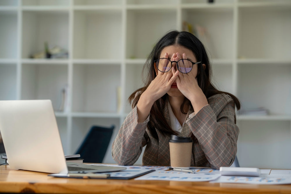 Stressed and tired woman sitting at a desk with a coffee, rubbing her eyes