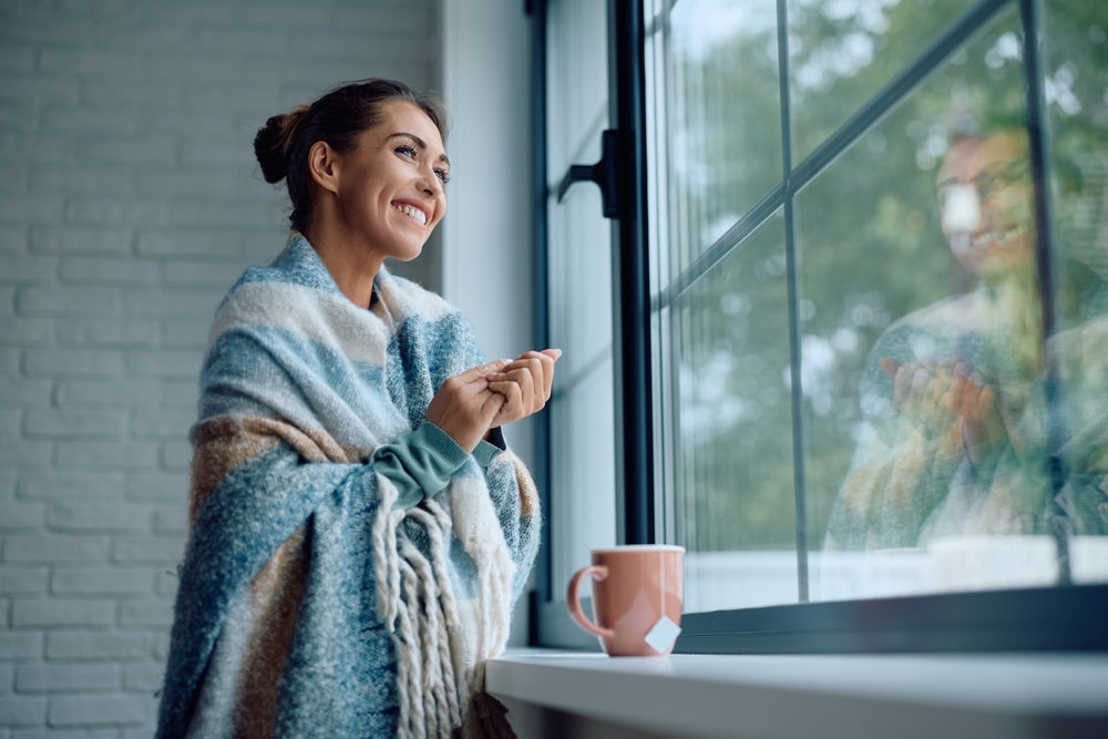 Woman smiling out of a bright window wrapped in a blanket looking well rested with a cup of tea